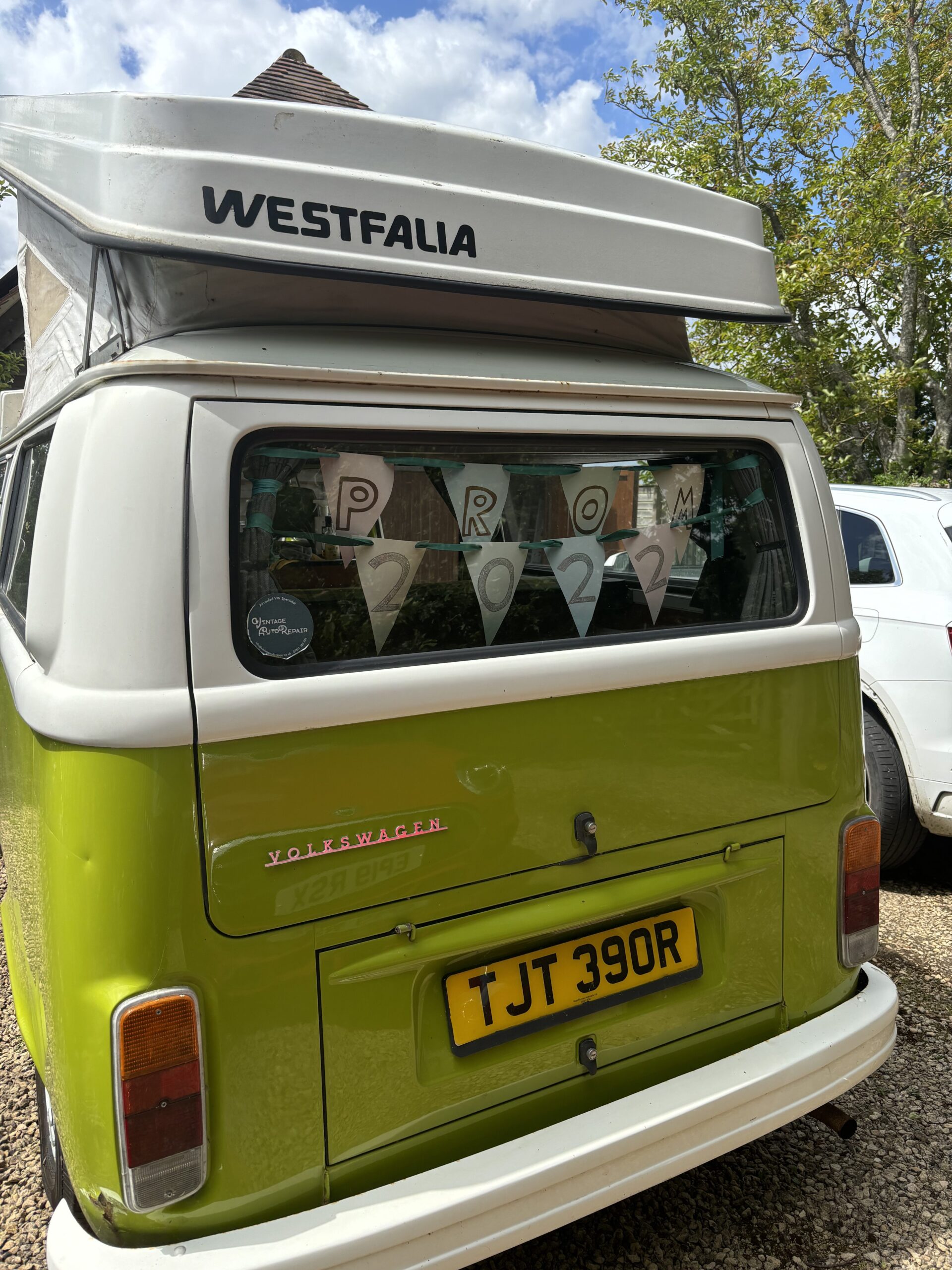 A green and white Volkswagen Westfalia camper van is parked outdoors. The rear window is decorated with triangular flags spelling 