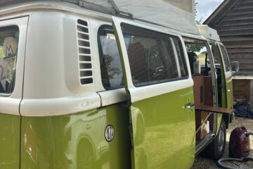 A green and white vintage Volkswagen camper van is parked with its side door open and an extended pop-up roof. Inside, wooden cabinetry is visible along with various camping equipment. The van is set on a gravel surface next to a wooden building, with a clear blue sky in the background.