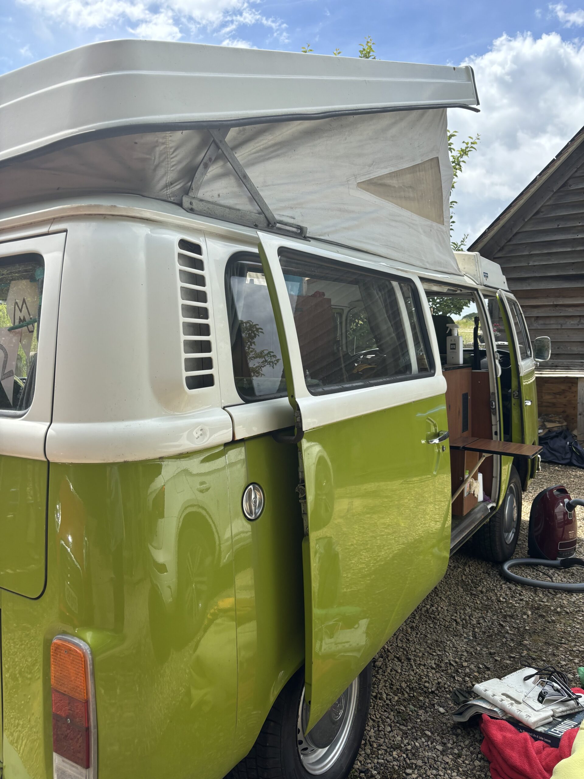 A green and white vintage Volkswagen camper van is parked with its side door open and an extended pop-up roof. Inside, wooden cabinetry is visible along with various camping equipment. The van is set on a gravel surface next to a wooden building, with a clear blue sky in the background.