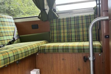 The image shows the interior of a vintage camper van. The seating area has green and yellow plaid fabric cushions. Green curtains frame the window, and the seats are built into a faux wood paneling structure. The floor is carpeted in green. A white electrical outlet is installed below the seat.