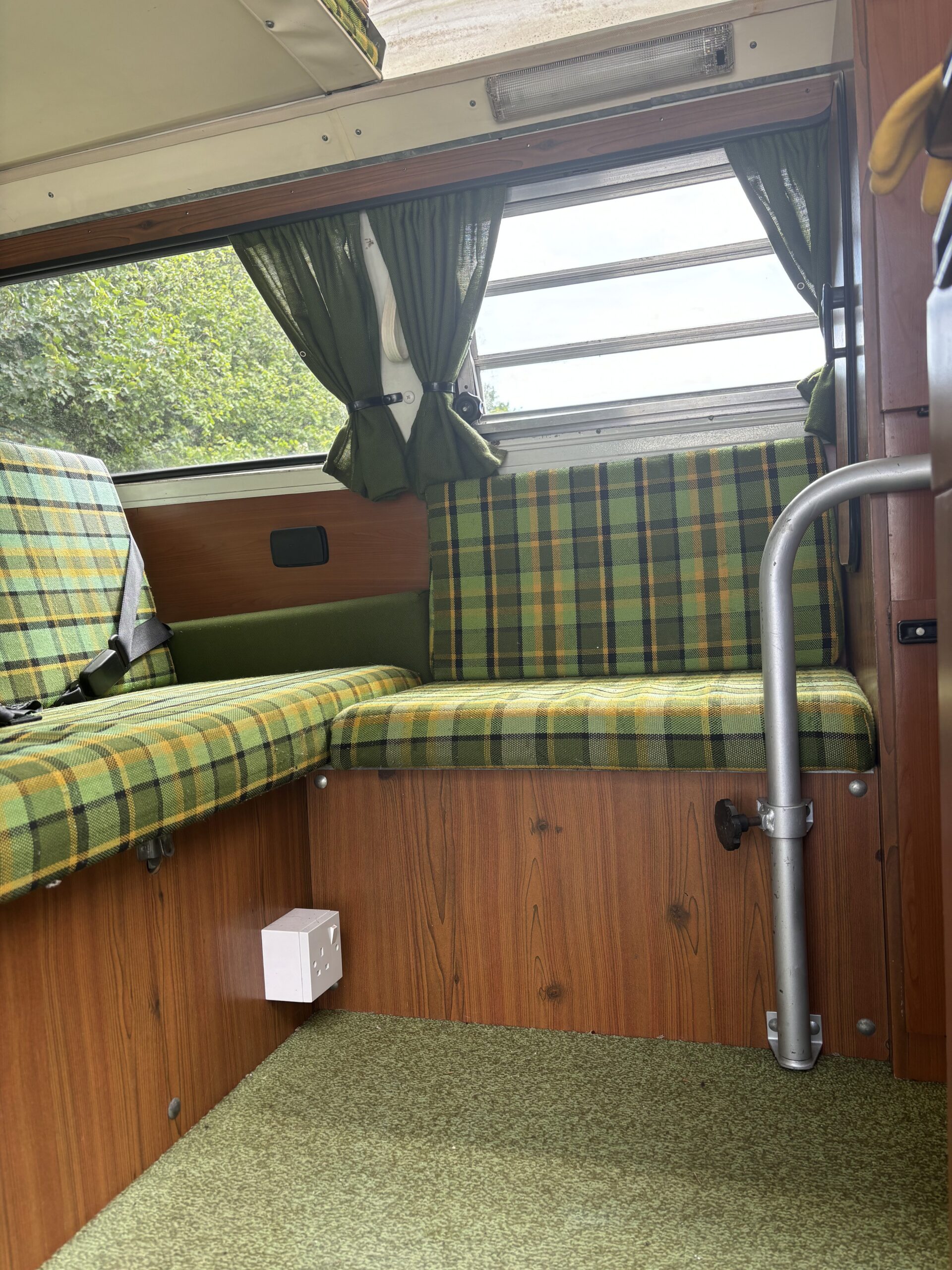 The image shows the interior of a vintage camper van. The seating area has green and yellow plaid fabric cushions. Green curtains frame the window, and the seats are built into a faux wood paneling structure. The floor is carpeted in green. A white electrical outlet is installed below the seat.