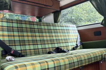 Interior view of a vintage camper van featuring a green and yellow plaid cushioned bench seat against wood-paneled walls. Two seat belts are fastened on the bench. The van also has green curtains on the windows, overhead wooden storage, and a power outlet is visible below the seat.