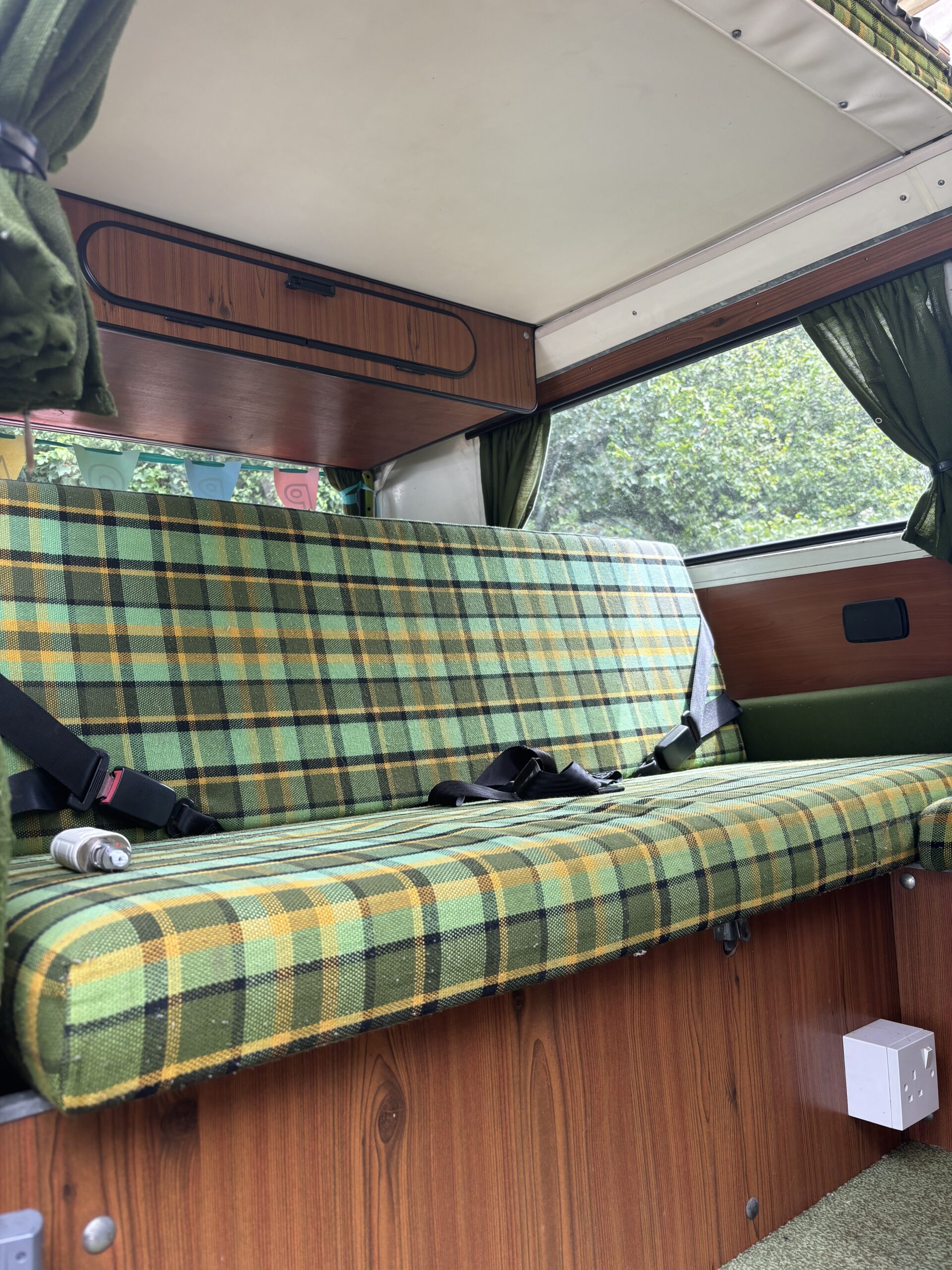 Interior view of a vintage camper van featuring a green and yellow plaid cushioned bench seat against wood-paneled walls. Two seat belts are fastened on the bench. The van also has green curtains on the windows, overhead wooden storage, and a power outlet is visible below the seat.