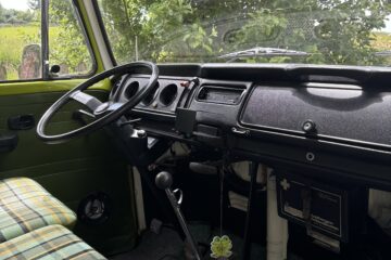 Interior of a vintage green-paneled van showing the dashboard, steering wheel, and manual gear shift. The seats are upholstered in a yellow and green plaid pattern. A first aid kit is visible under the dashboard, and a tree with green foliage can be seen through the windows.