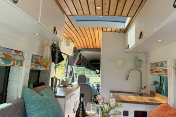 A cozy camper van interior features a wooden ceiling with recessed lighting, a skylight, a gray couch with colorful pillows, a compact kitchen with a sink, and a rustic wooden table displaying a wine bottle, glasses, and a mint vase with flowers. The windows on both sides are draped with vibrant curtains.