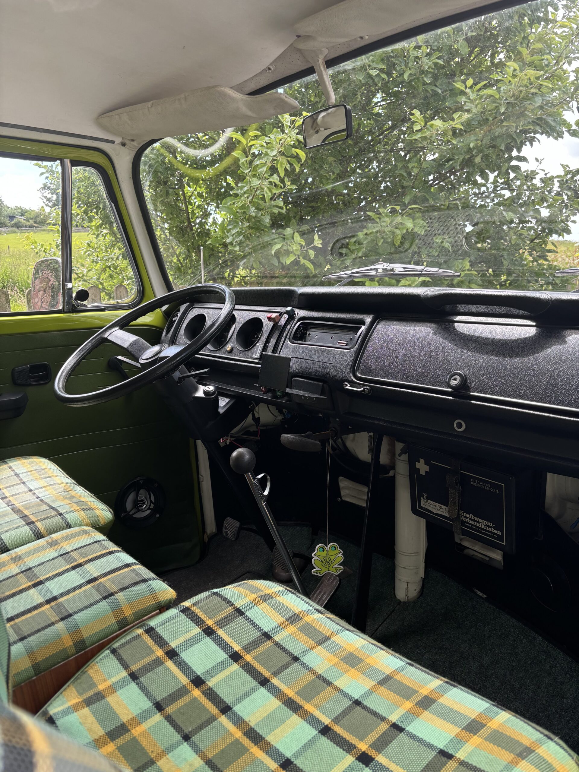 Interior of a vintage green-paneled van showing the dashboard, steering wheel, and manual gear shift. The seats are upholstered in a yellow and green plaid pattern. A first aid kit is visible under the dashboard, and a tree with green foliage can be seen through the windows.