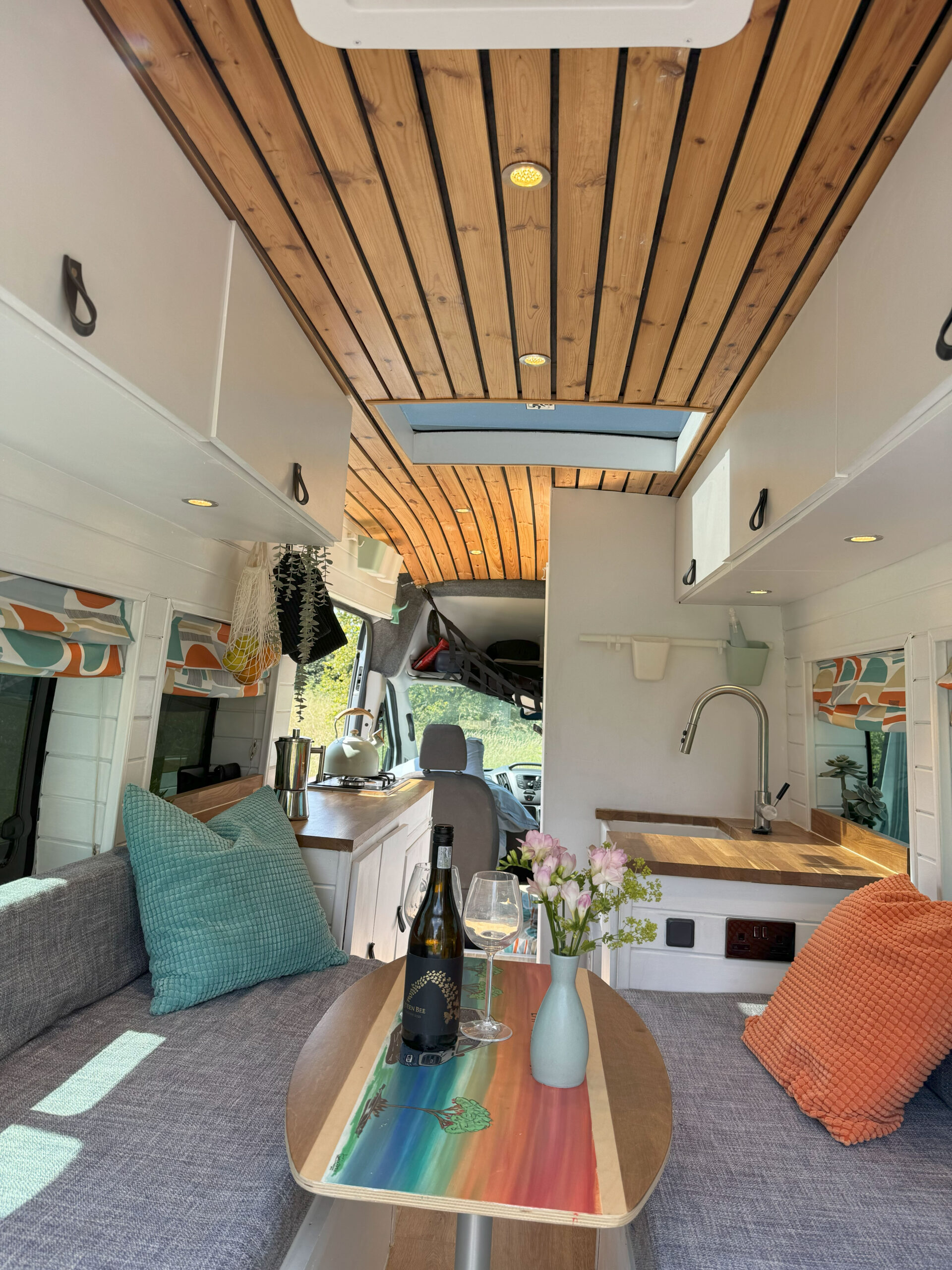 A cozy camper van interior features a wooden ceiling with recessed lighting, a skylight, a gray couch with colorful pillows, a compact kitchen with a sink, and a rustic wooden table displaying a wine bottle, glasses, and a mint vase with flowers. The windows on both sides are draped with vibrant curtains.