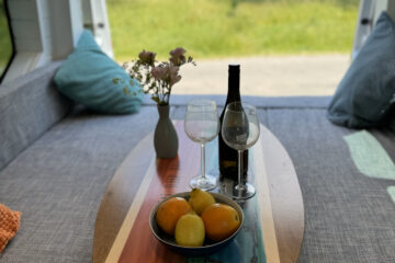 The interior of a camper van with a colorful oval wooden table adorned with two wine glasses, a bottle of wine, and a bowl of fruit. There are two cushioned benches, a vase with flowers, and a scenic view of a grassy field through the open back door.