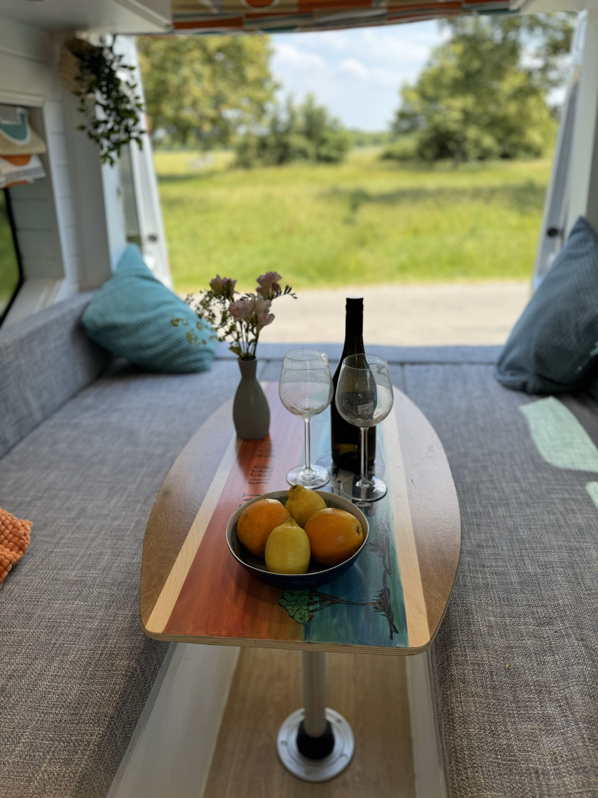 The interior of a camper van with a colorful oval wooden table adorned with two wine glasses, a bottle of wine, and a bowl of fruit. There are two cushioned benches, a vase with flowers, and a scenic view of a grassy field through the open back door.