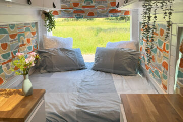 A cozy van interior features a bed with gray sheets and two pillows, nestled against a wall with colorful, geometric-patterned curtains. Above the bed, small potted plants hang for decoration. The ceiling is wooden, and there are white cabinets with wood countertops on either side.