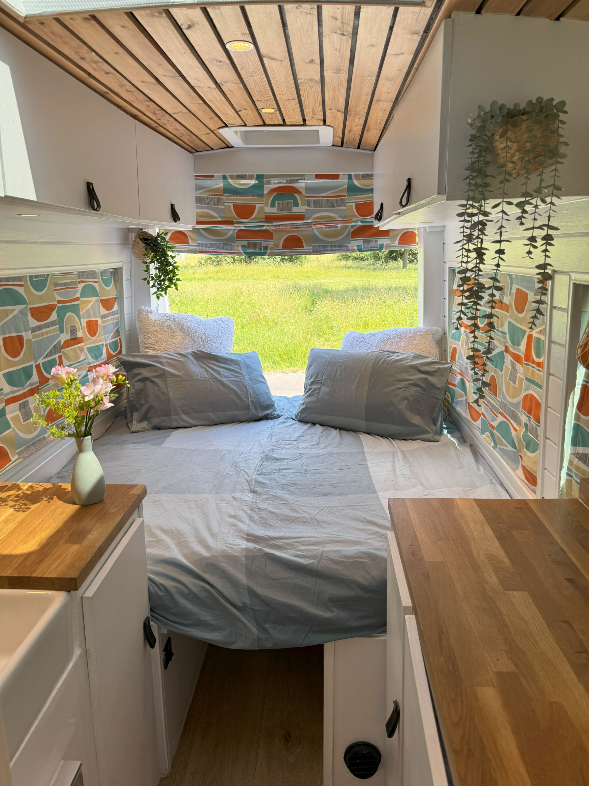 A cozy van interior features a bed with gray sheets and two pillows, nestled against a wall with colorful, geometric-patterned curtains. Above the bed, small potted plants hang for decoration. The ceiling is wooden, and there are white cabinets with wood countertops on either side.