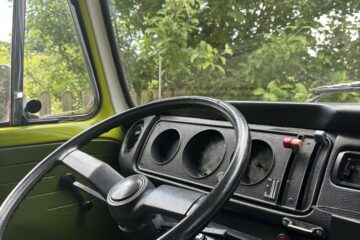 Interior view of a vintage vehicle showing a minimalist black steering wheel, simple dashboard with three round gauges, and a few control switches. The window reveals green foliage outside, suggesting a scenic, natural setting. The interior door panel is olive green with a manual window crank.