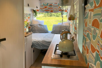 A cozy, brightly lit camper van interior features a small kitchen area on the right with a kettle on the stove, storage below, and colorful patterned curtains. At the rear, a bed with grey bedding and green and orange pillows sits surrounded by windows with sheer blinds. Plants hang from the ceiling.