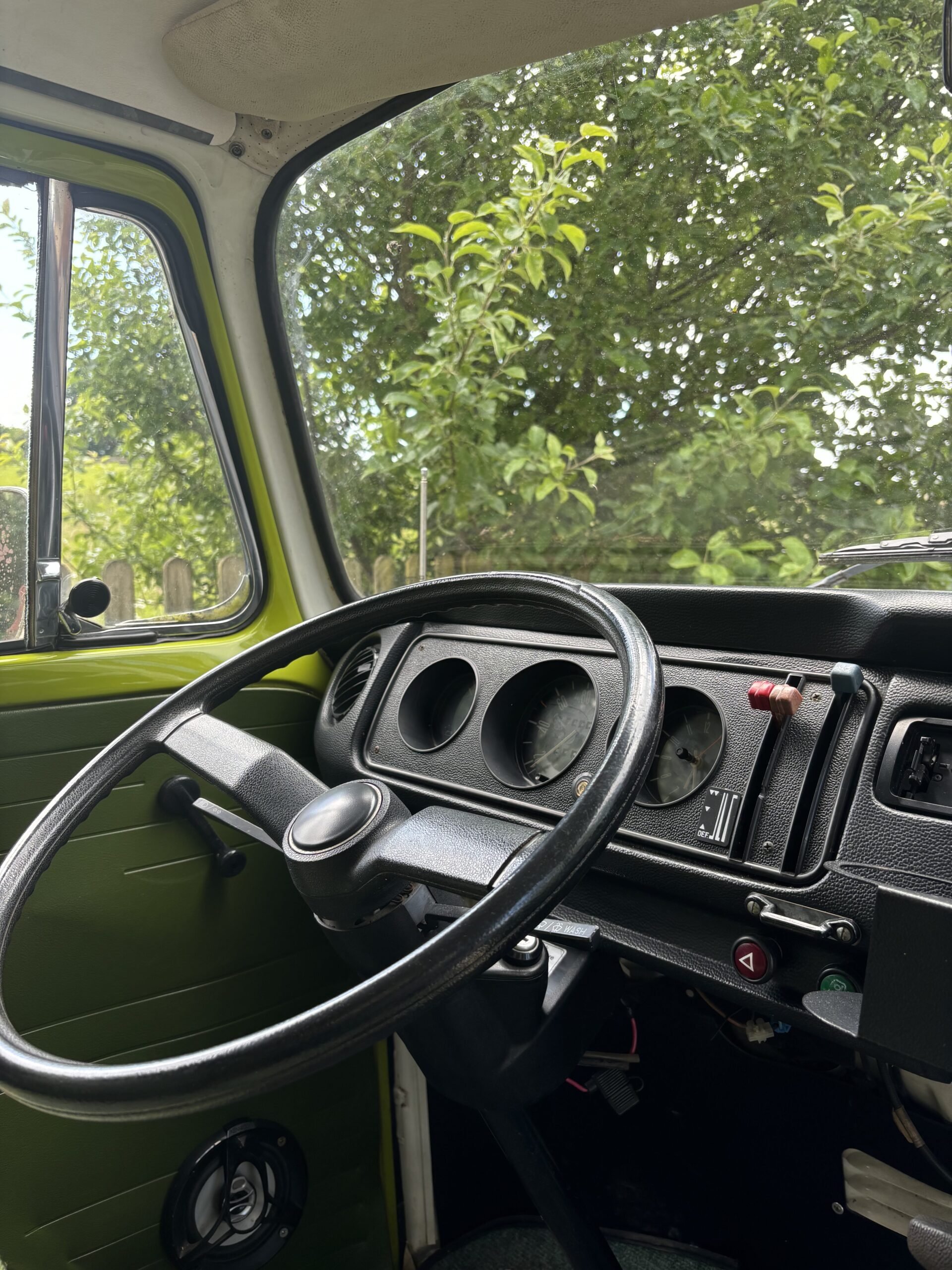 Interior view of a vintage vehicle showing a minimalist black steering wheel, simple dashboard with three round gauges, and a few control switches. The window reveals green foliage outside, suggesting a scenic, natural setting. The interior door panel is olive green with a manual window crank.