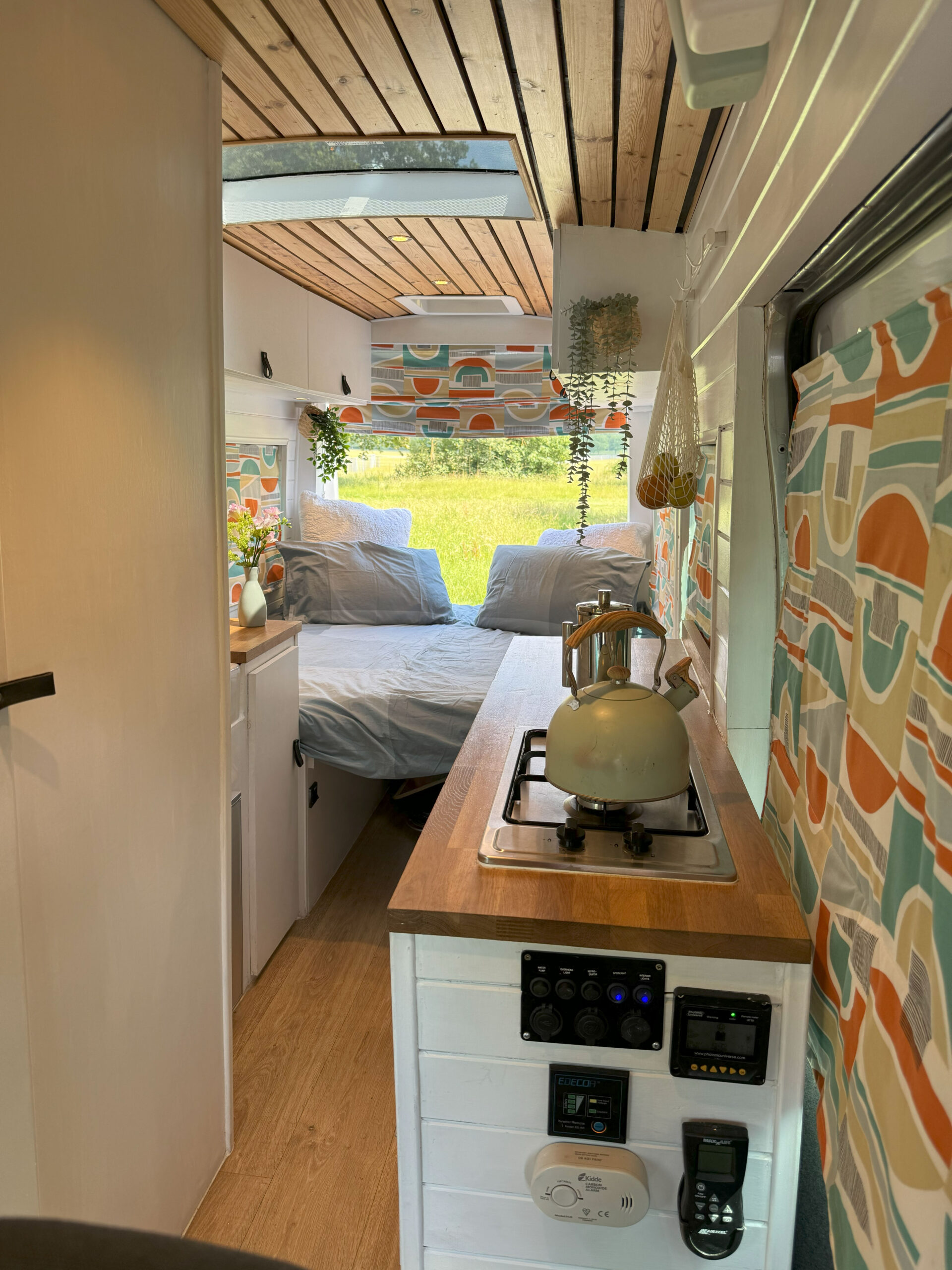 A cozy, brightly lit camper van interior features a small kitchen area on the right with a kettle on the stove, storage below, and colorful patterned curtains. At the rear, a bed with grey bedding and green and orange pillows sits surrounded by windows with sheer blinds. Plants hang from the ceiling.