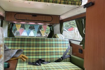 Interior of a retro camper van with a green and yellow checkered bench seat that has seat belts. The ceiling is curved, with a white upper section. Colorful triangular bunting hangs across the back window. There's a white electrical outlet near a small step. Curtains frame the windows.