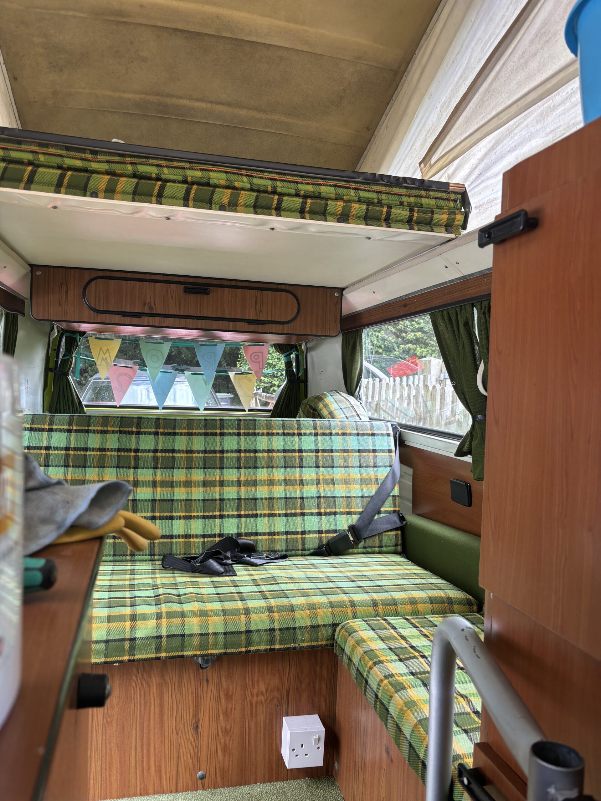 Interior of a retro camper van with a green and yellow checkered bench seat that has seat belts. The ceiling is curved, with a white upper section. Colorful triangular bunting hangs across the back window. There's a white electrical outlet near a small step. Curtains frame the windows.