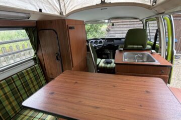 Interior view of a vintage camper van with a wood-finished table in the center. Green plaid seating is on the left and front, with a kitchenette featuring a sink on the right. A person's legs and shoes are visible in the foreground. The pop-top ceiling is raised, revealing a mesh window.