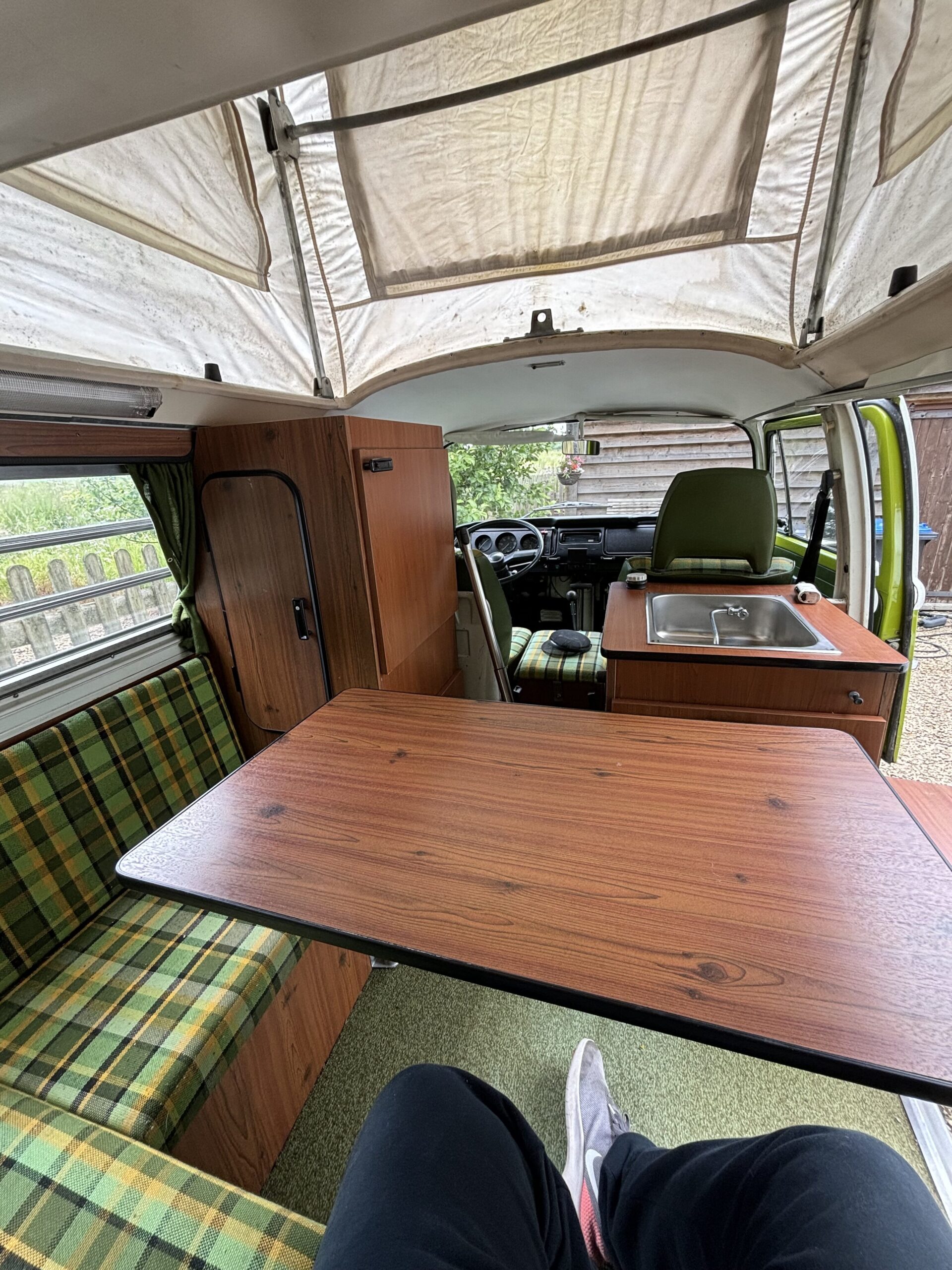 Interior view of a vintage camper van with a wood-finished table in the center. Green plaid seating is on the left and front, with a kitchenette featuring a sink on the right. A person's legs and shoes are visible in the foreground. The pop-top ceiling is raised, revealing a mesh window.