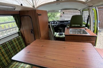 Interior view of a vintage camper van with predominantly green plaid upholstery. It features a fold-out wooden table, a small kitchen area with a sink, and storage cabinets. The roof is partially raised for extra headroom, and a partially open door reveals a wooden fence and greenery outside.