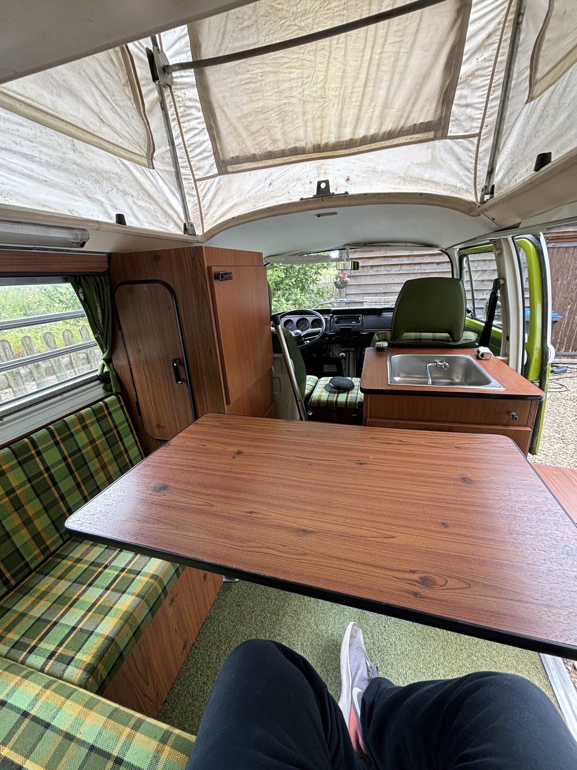 Interior view of a vintage camper van with predominantly green plaid upholstery. It features a fold-out wooden table, a small kitchen area with a sink, and storage cabinets. The roof is partially raised for extra headroom, and a partially open door reveals a wooden fence and greenery outside.