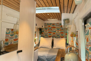 Interior of a compact, stylish camper van with wooden ceiling and bright decor. A cozy double bed with white bedding is set up in the back, bordered by colorful patterned curtains. The kitchenette includes a stove with a blue kettle, and there's a wooden countertop with modern fixtures.