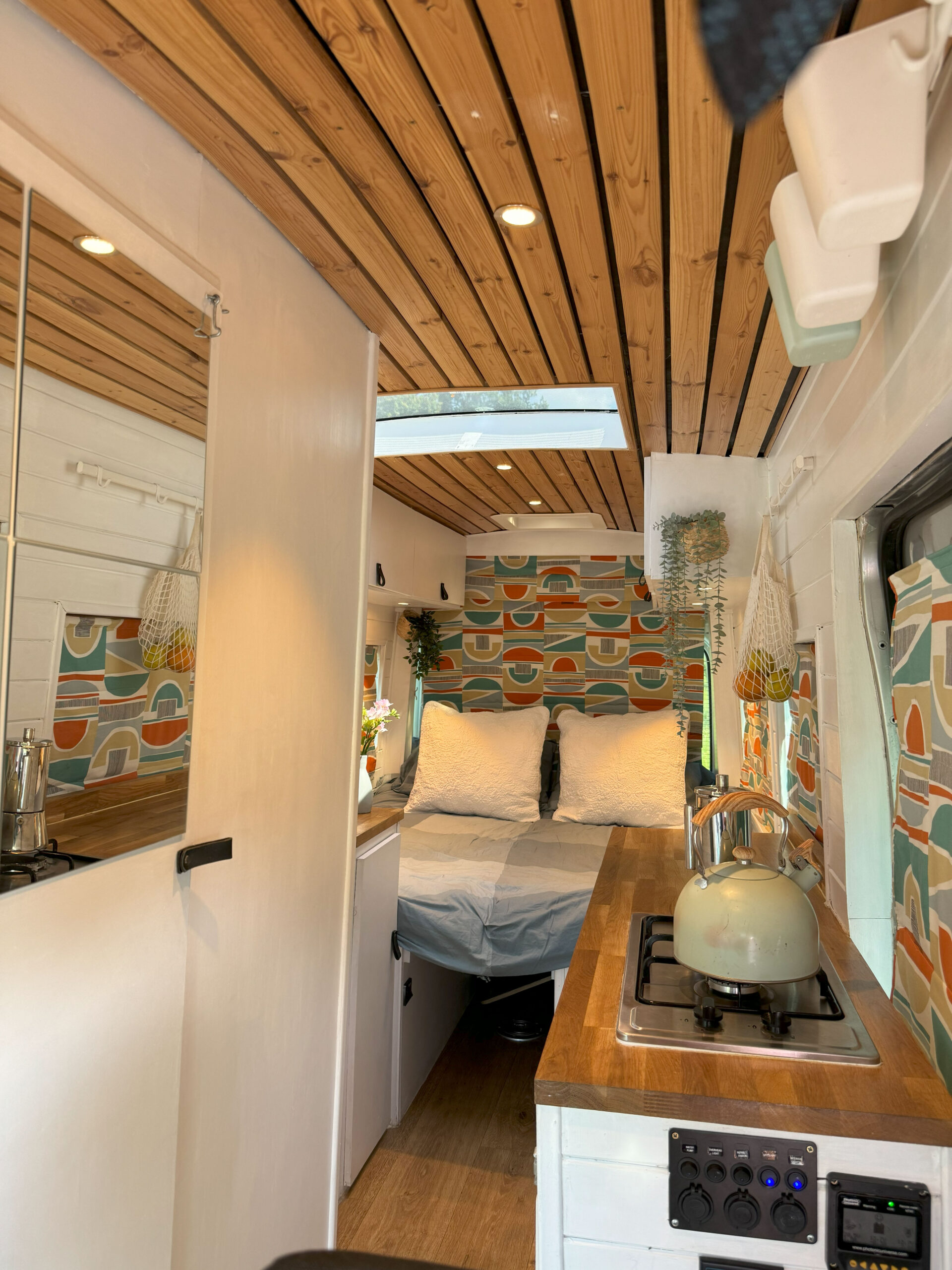 Interior of a compact, stylish camper van with wooden ceiling and bright decor. A cozy double bed with white bedding is set up in the back, bordered by colorful patterned curtains. The kitchenette includes a stove with a blue kettle, and there's a wooden countertop with modern fixtures.