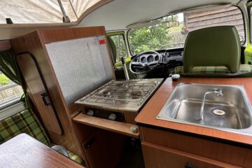 Interior of a vintage camper van with a pop-up roof. The foreground shows a wooden table with a plaid seat, a gas stove, and a metal sink installed in wooden cabinetry. The driver's area is visible with the green driver’s seat facing the steering wheel. A window offers an outside view.