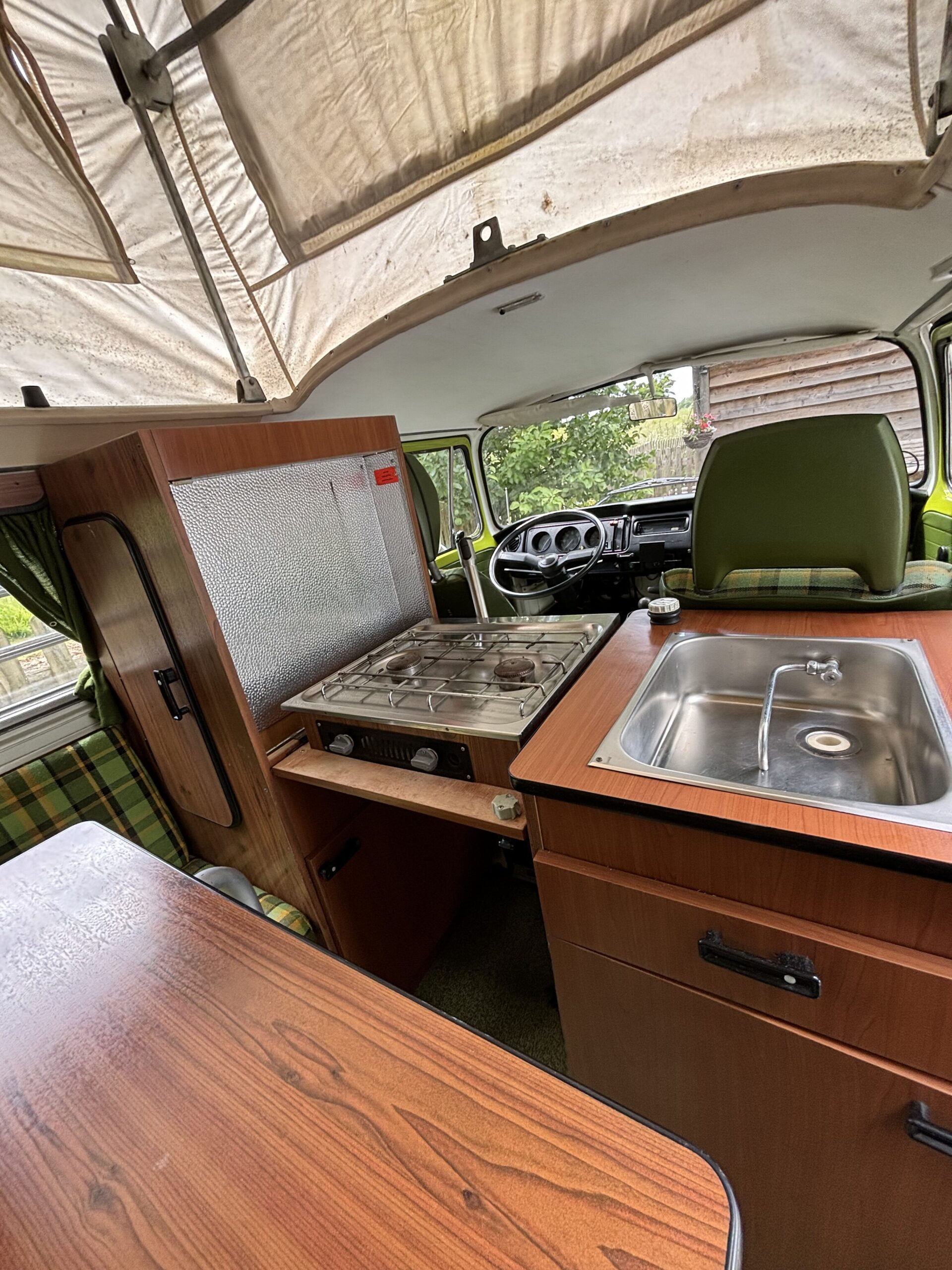 Interior of a vintage camper van with a pop-up roof. The foreground shows a wooden table with a plaid seat, a gas stove, and a metal sink installed in wooden cabinetry. The driver's area is visible with the green driver’s seat facing the steering wheel. A window offers an outside view.