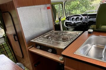 Interior of a vintage camper van featuring a compact kitchen with a small stainless steel sink, a two-burner gas stove, and a wooden countertop. Adjacent is a vertical fridge with a textured metallic surface. The driver's area is visible in the background, with green seats.