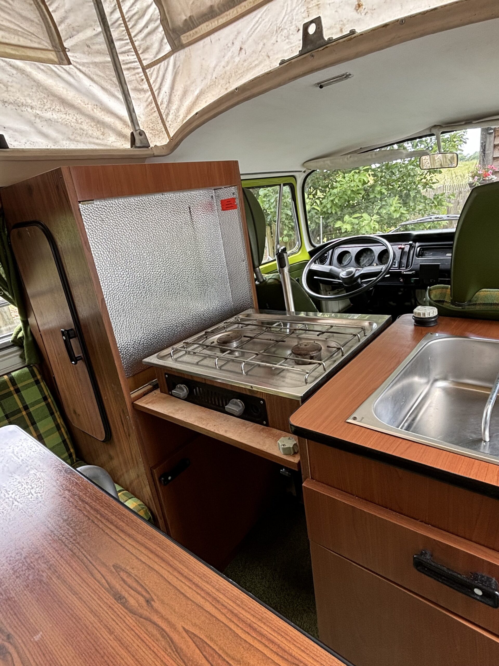 Interior of a vintage camper van featuring a compact kitchen with a small stainless steel sink, a two-burner gas stove, and a wooden countertop. Adjacent is a vertical fridge with a textured metallic surface. The driver's area is visible in the background, with green seats.