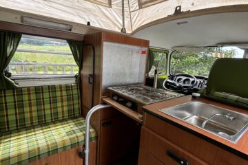 Interior of a vintage camper van with a cozy setup. Features include a stove and sink on the right, a green plaid cushioned bench seat on the left, and wood paneling throughout. Curtains frame the window behind the bench, and the elevated, tent-like roof reveals canvas siding and supports.