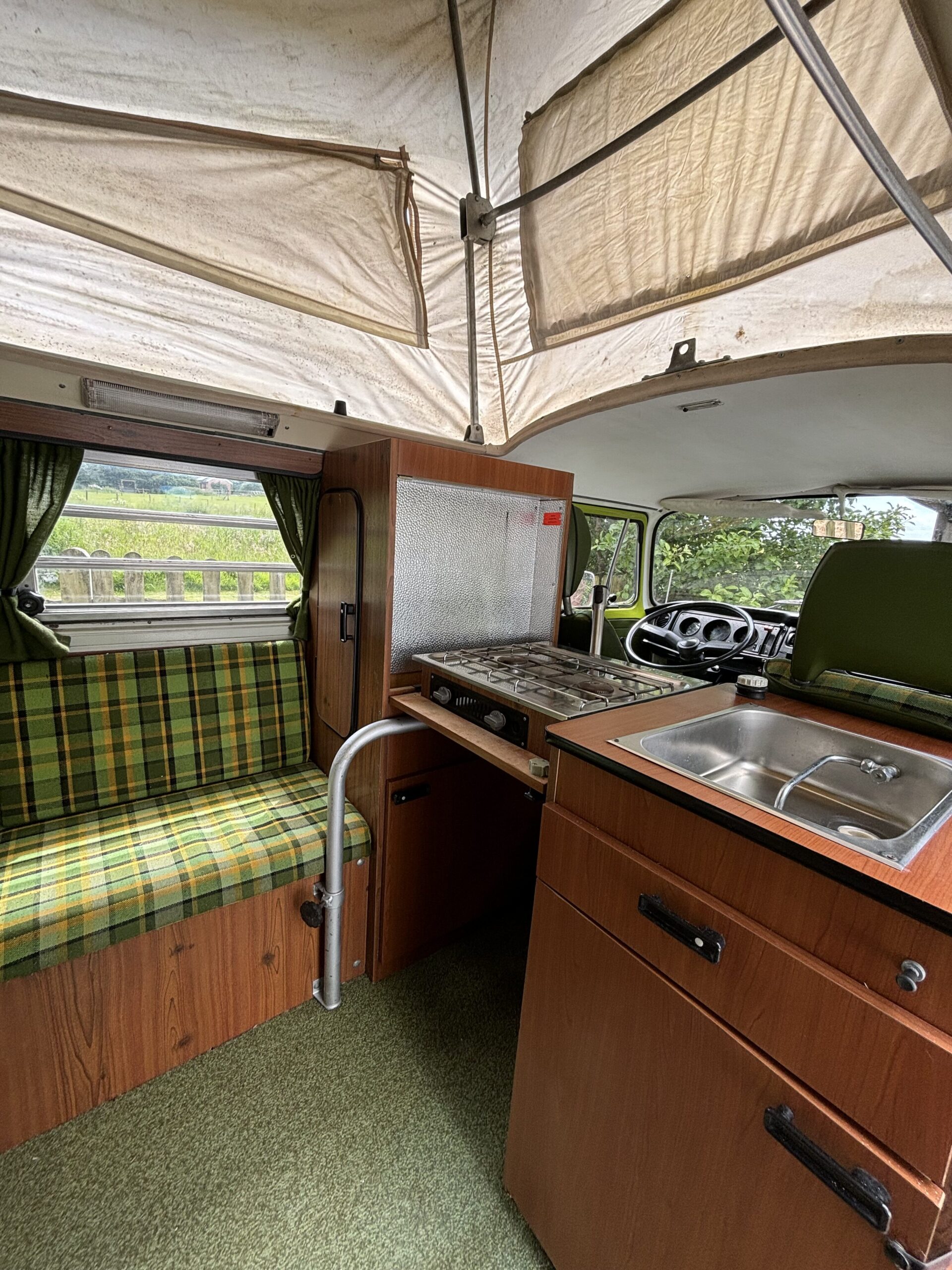 Interior of a vintage camper van with a cozy setup. Features include a stove and sink on the right, a green plaid cushioned bench seat on the left, and wood paneling throughout. Curtains frame the window behind the bench, and the elevated, tent-like roof reveals canvas siding and supports.