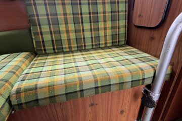 A plaid-patterned seat with green, yellow, and orange tones in a small, wooden-paneled space, possibly a vintage camper or caravan. The seat cushion extends along the wall below a window with green curtains. A curved metal frame is visible, attached to the wooden paneling.