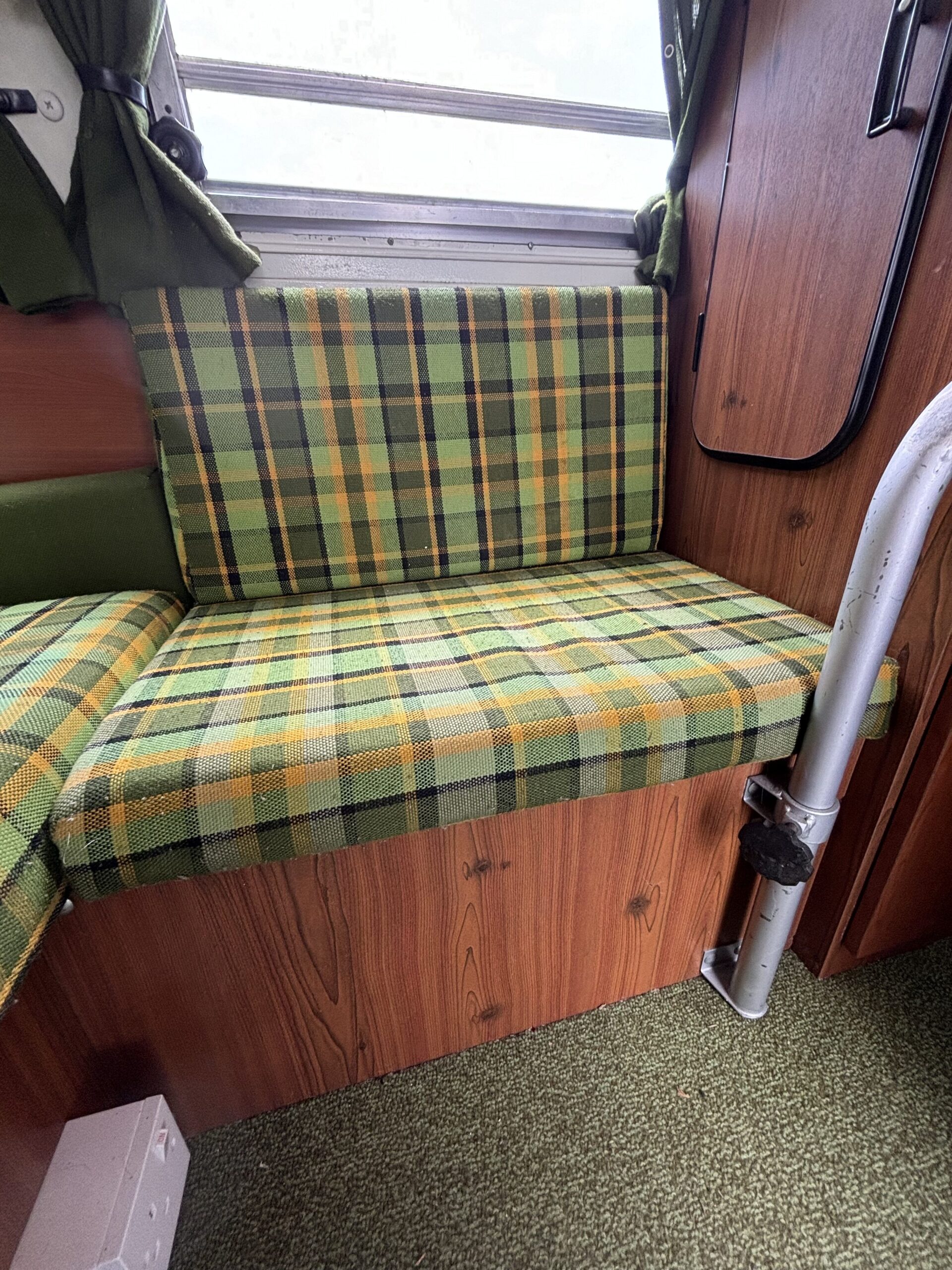 A plaid-patterned seat with green, yellow, and orange tones in a small, wooden-paneled space, possibly a vintage camper or caravan. The seat cushion extends along the wall below a window with green curtains. A curved metal frame is visible, attached to the wooden paneling.