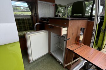 A retro green and white camper van with its side door open reveals the interior. The setup includes a small wooden kitchenette featuring a mini-fridge, a stove, and a cabinet. There's also a fold-out table with a bottle. In the back, green plaid seating is partly visible.