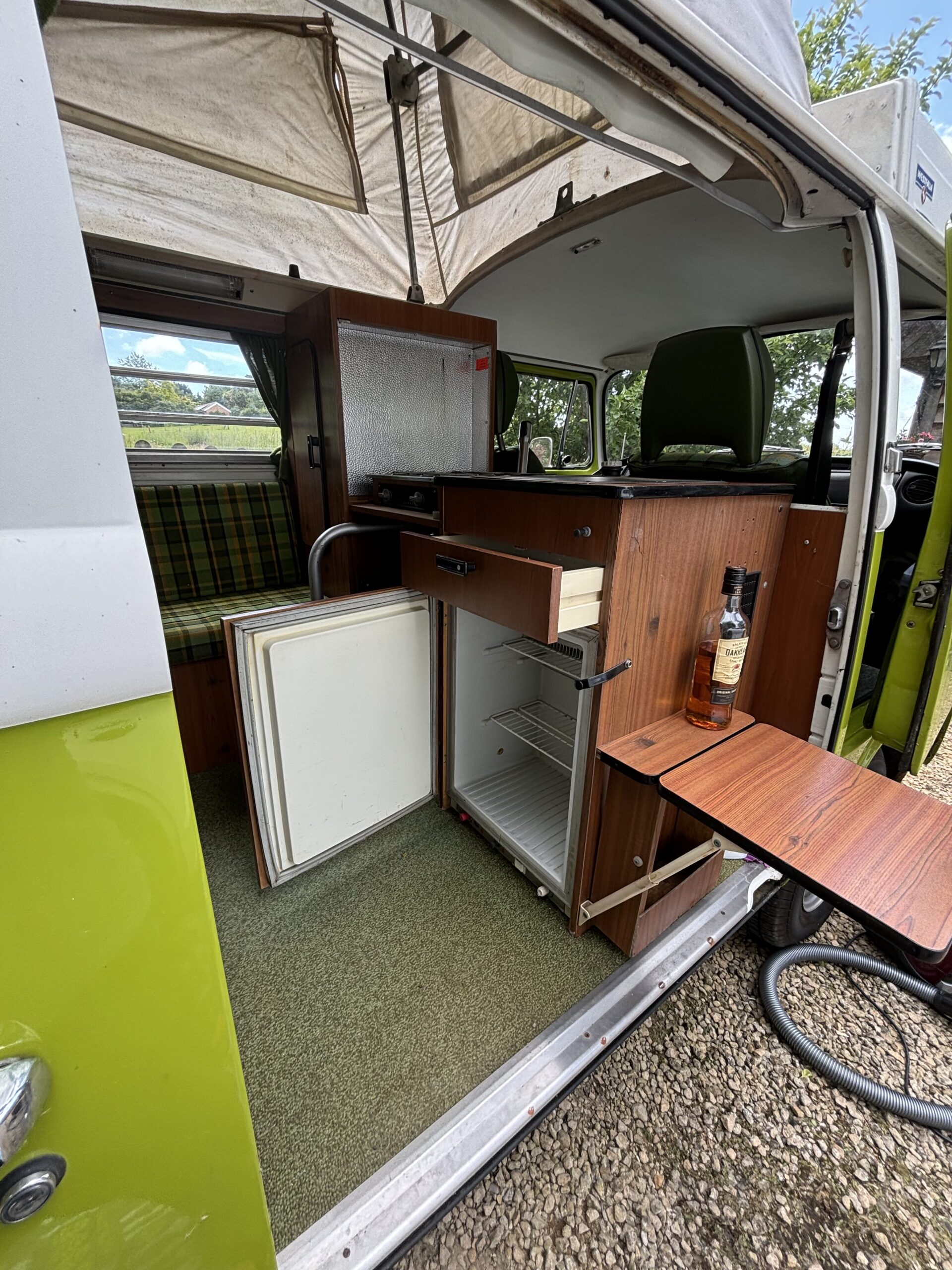 A retro green and white camper van with its side door open reveals the interior. The setup includes a small wooden kitchenette featuring a mini-fridge, a stove, and a cabinet. There's also a fold-out table with a bottle. In the back, green plaid seating is partly visible.