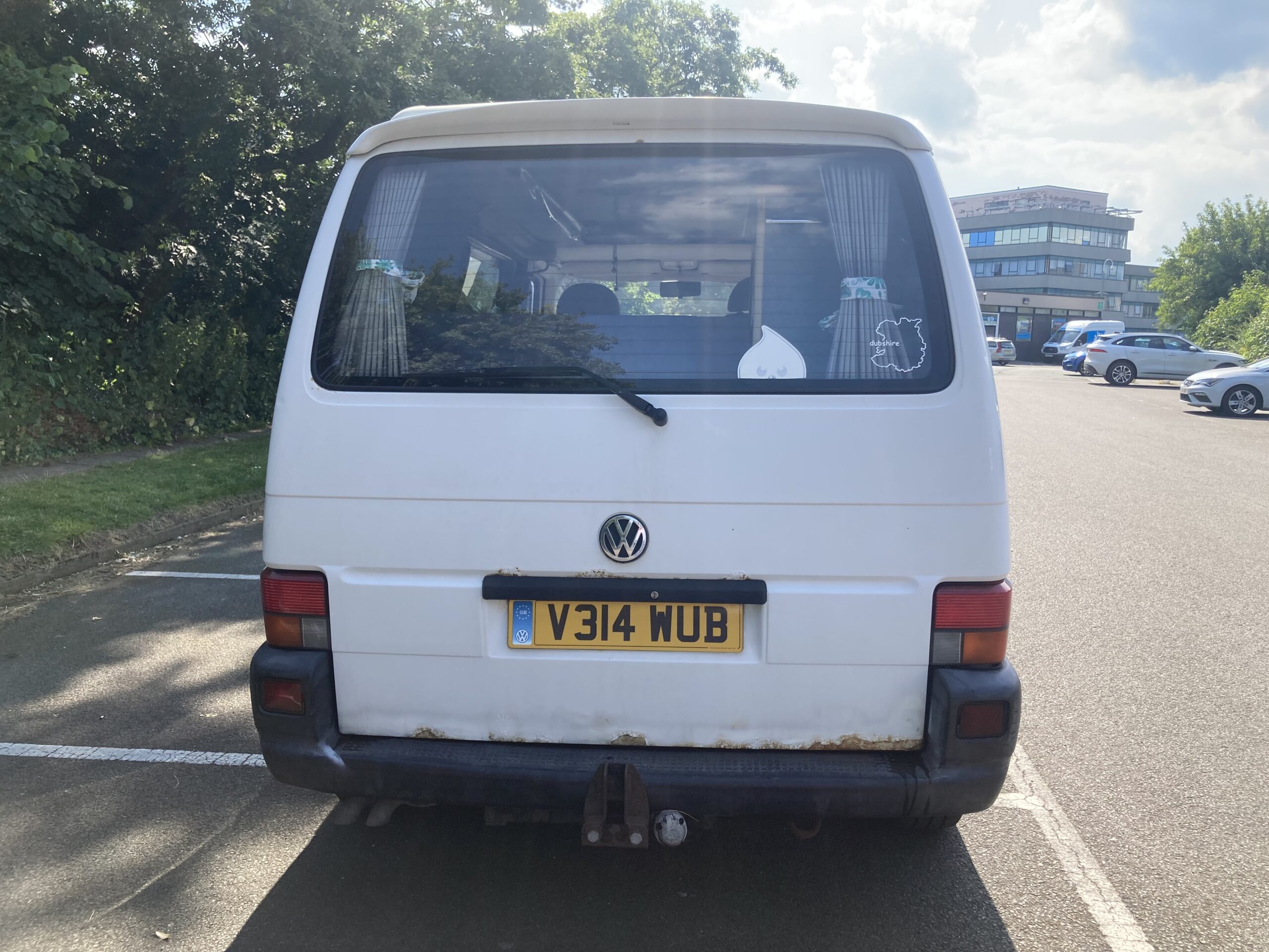 A white Volkswagen van with a UK license plate (V314 WUB) is parked on an asphalt lot. The van has visible rust on the rear bumper and stickers on its rear window. Trees line the left side, and a multi-story building is in the background on the right. The sky is partly cloudy.