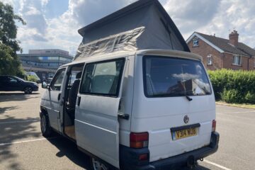 A white Volkswagen camper van with the pop-up roof extended is parked in a lot under a partly cloudy sky. The van's sliding door is open, revealing part of the interior. A few buildings and a lush green hedge appear in the background. The vehicle's license plate reads "V314 WUB.