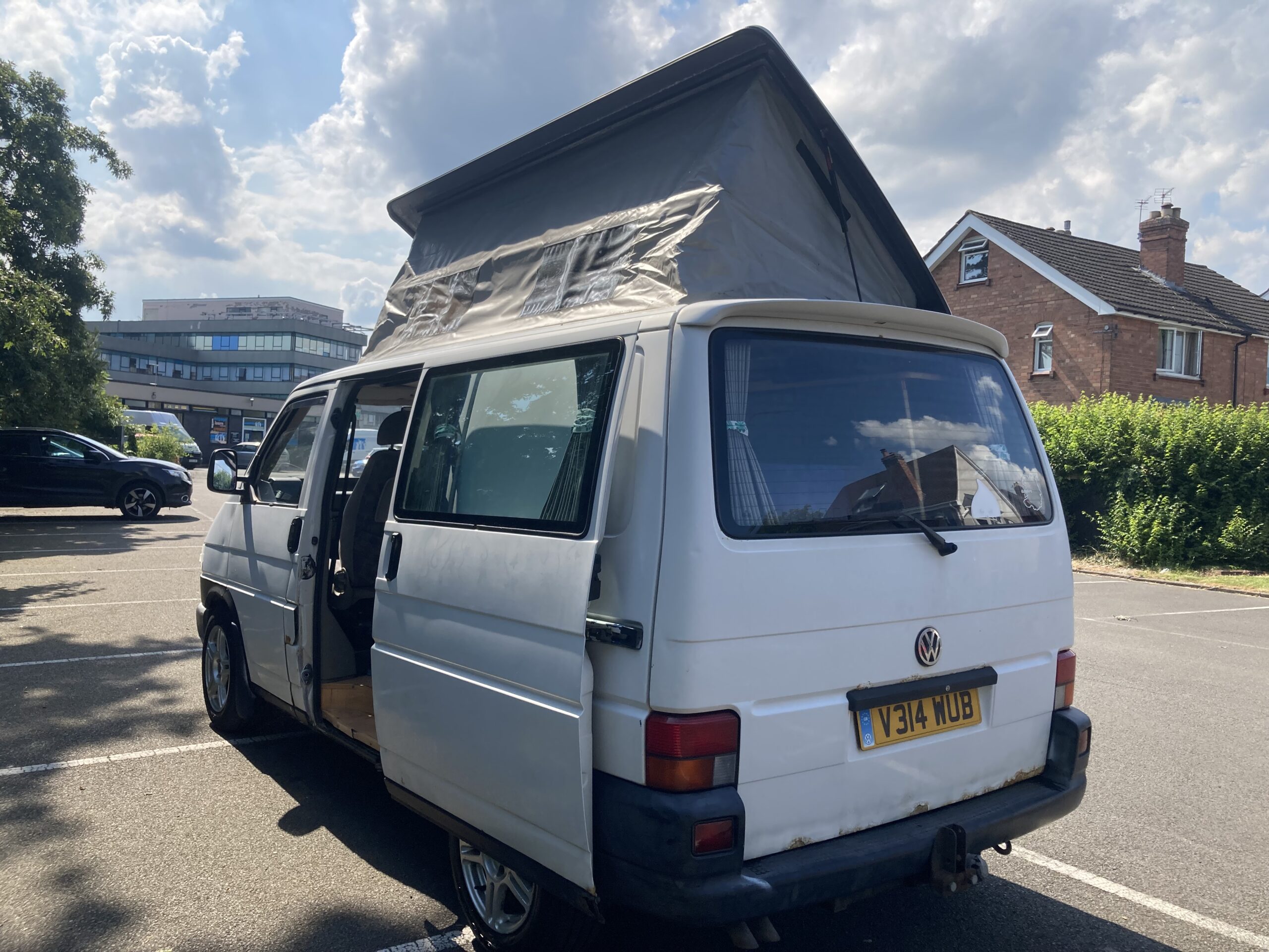 A white Volkswagen camper van with the pop-up roof extended is parked in a lot under a partly cloudy sky. The van's sliding door is open, revealing part of the interior. A few buildings and a lush green hedge appear in the background. The vehicle's license plate reads