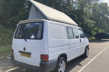 A white camper van with an extended roof is parked in a parking lot next to a forested area. Its license plate reads "V314 WUB" and it has alloy wheels. The van has a slightly opened window on the side and stickers on the rear windows. It is a sunny day with clear skies and surrounding greenery.
