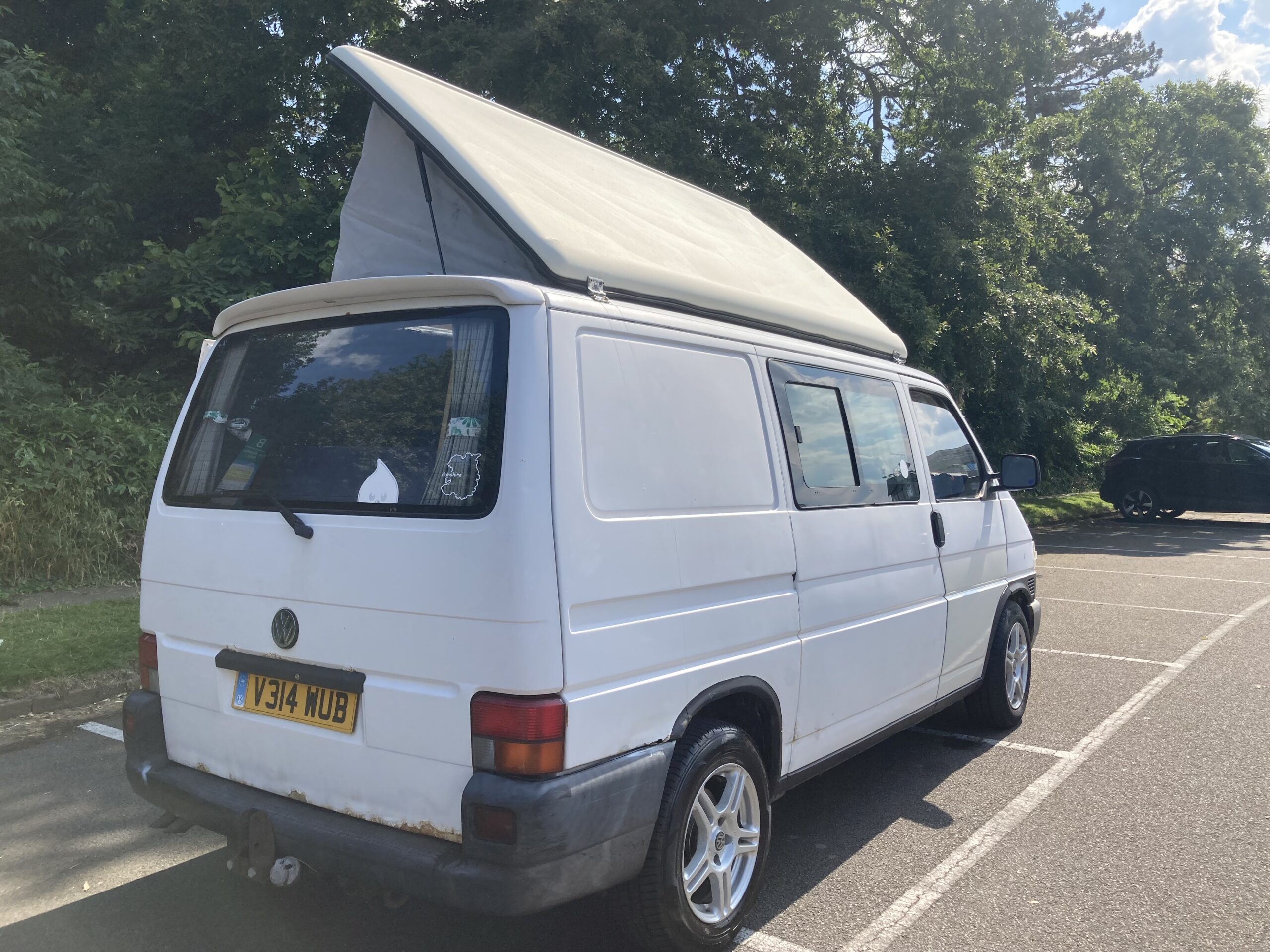 A white camper van with an extended roof is parked in a parking lot next to a forested area. Its license plate reads