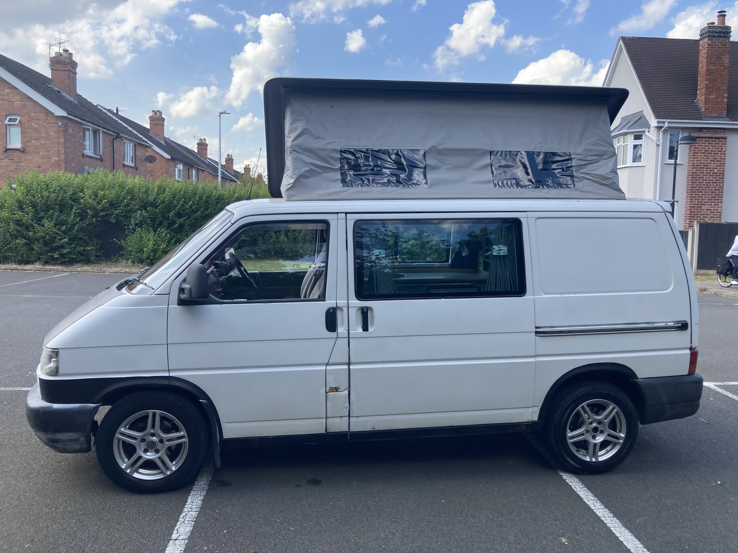A white camper van with a pop-up roof is parked in an asphalt parking lot. The roof is elevated, with canvas sides and plastic windows. The van has alloy wheels and is set against a suburban backdrop with brick houses, green hedges, and a partly cloudy blue sky.