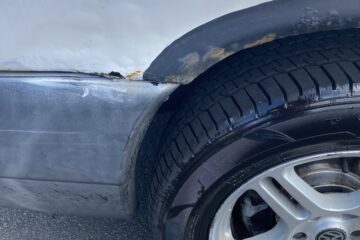Close-up of a car's wheel and part of the body. The car's white paint is damaged and shows rust along the wheel well. The tire and silver alloy wheel rim appear new and in good condition, contrasting with the worn appearance of the car's body. The car is parked on a paved surface.