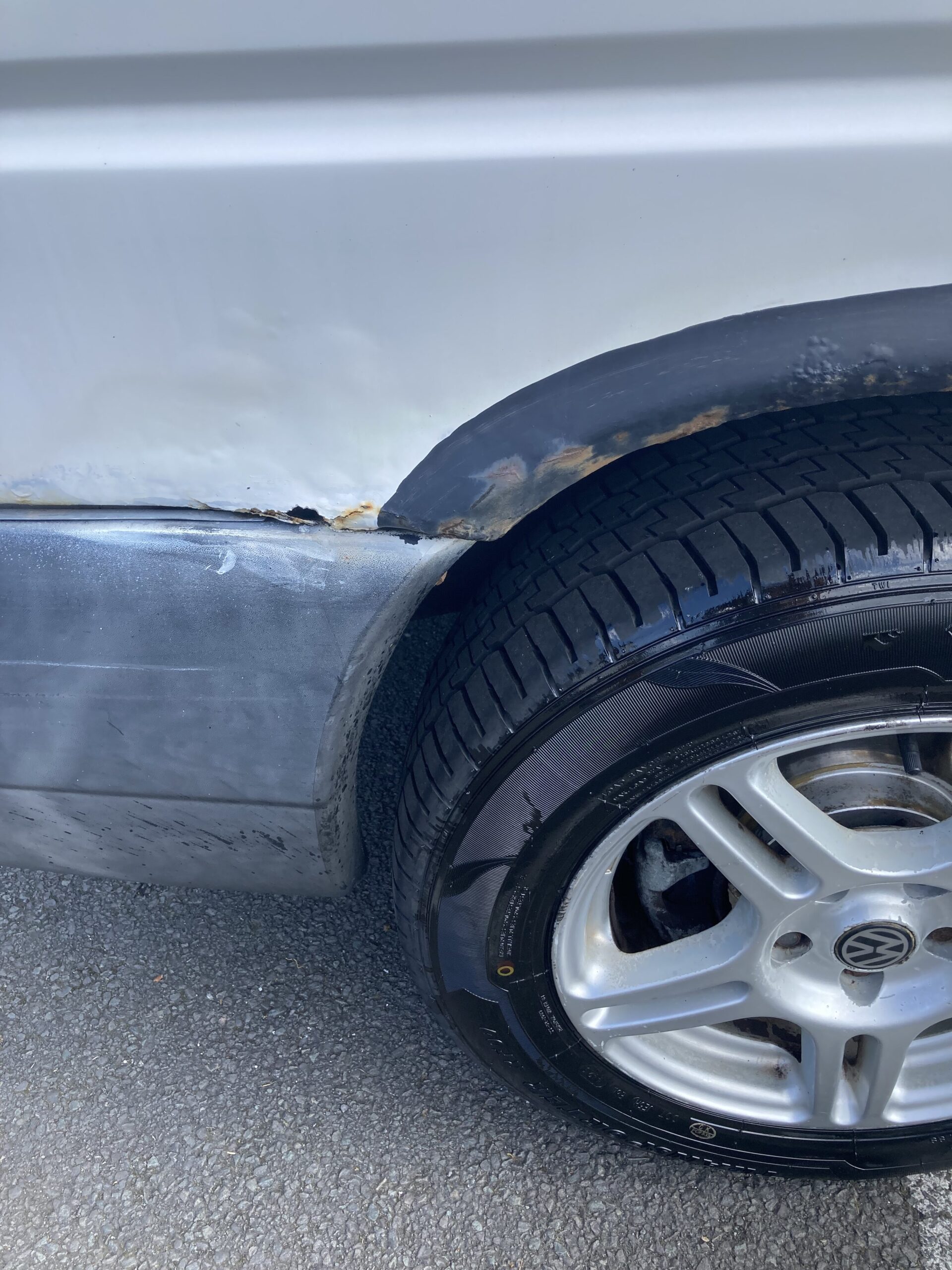 Close-up of a car's wheel and part of the body. The car's white paint is damaged and shows rust along the wheel well. The tire and silver alloy wheel rim appear new and in good condition, contrasting with the worn appearance of the car's body. The car is parked on a paved surface.