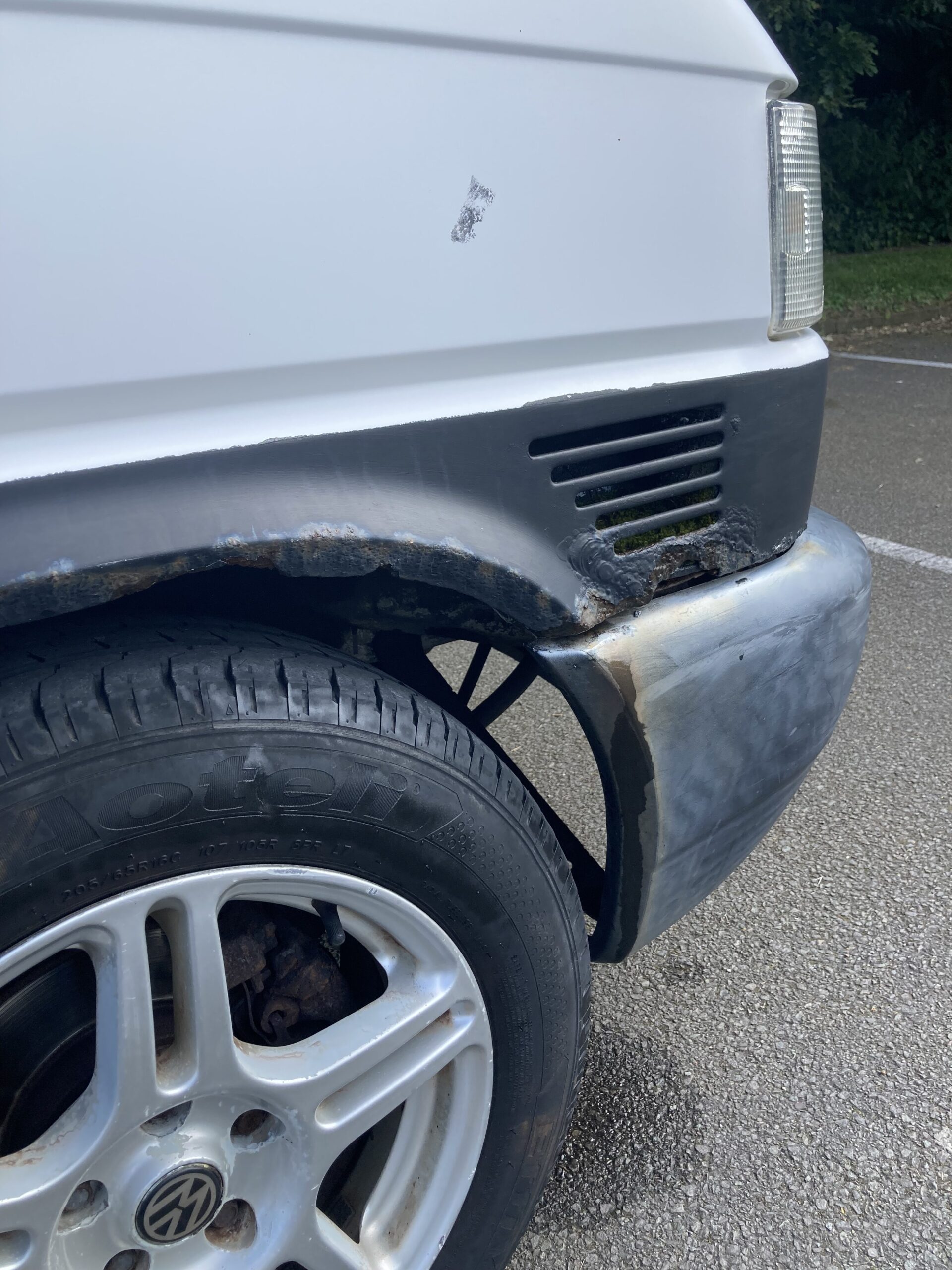 A close-up of a white vehicle's front right corner, showing rust and damage along the fender and bumper. The black trim is visibly deteriorated, exposing rust beneath. The tire and wheel are partly visible, and the car is parked on a concrete surface with greenery in the background.