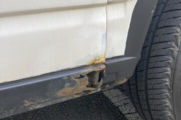 A close-up view of a white vehicle's lower side panel and wheel well. There is significant rust damage and holes near the bottom edge, with visible corrosion spreading upwards. The adjacent black trim and nearby tire appear in good condition. The car is parked on an asphalt surface with painted lines.