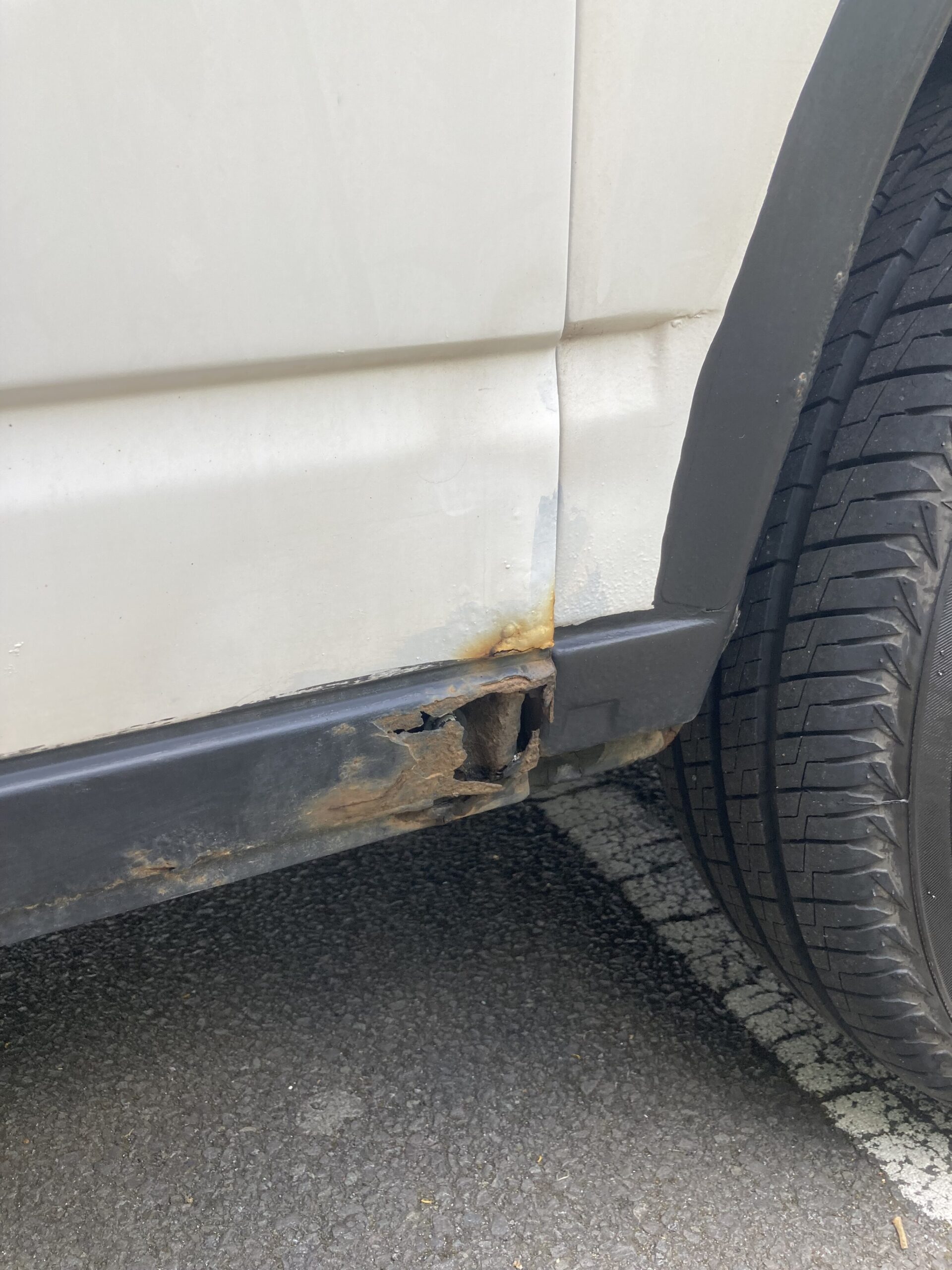 A close-up view of a white vehicle's lower side panel and wheel well. There is significant rust damage and holes near the bottom edge, with visible corrosion spreading upwards. The adjacent black trim and nearby tire appear in good condition. The car is parked on an asphalt surface with painted lines.