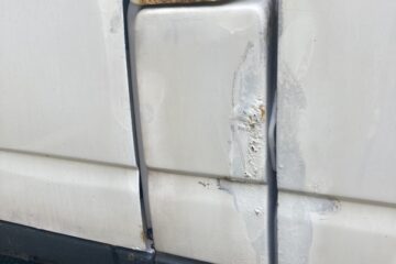 Close-up of a white vehicle's side with visible damage. The door seam shows significant rust, especially around a prominent hole near the middle of the door. Scratches and rust patches are also visible below the hole, indicating wear and tear. The vehicle is parked on a dark asphalt surface.