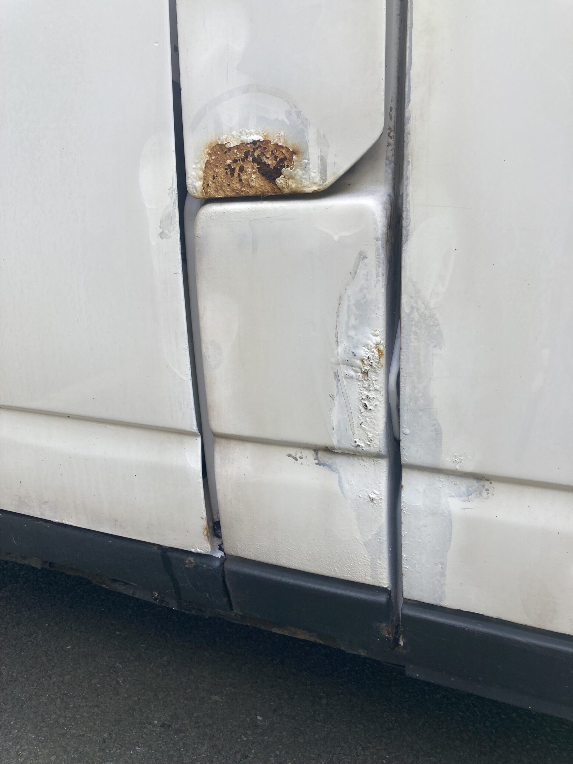 Close-up of a white vehicle's side with visible damage. The door seam shows significant rust, especially around a prominent hole near the middle of the door. Scratches and rust patches are also visible below the hole, indicating wear and tear. The vehicle is parked on a dark asphalt surface.